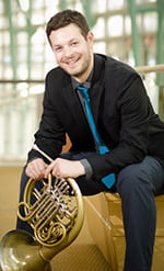 Josh Michal sitting with french horn, smiling, indoors